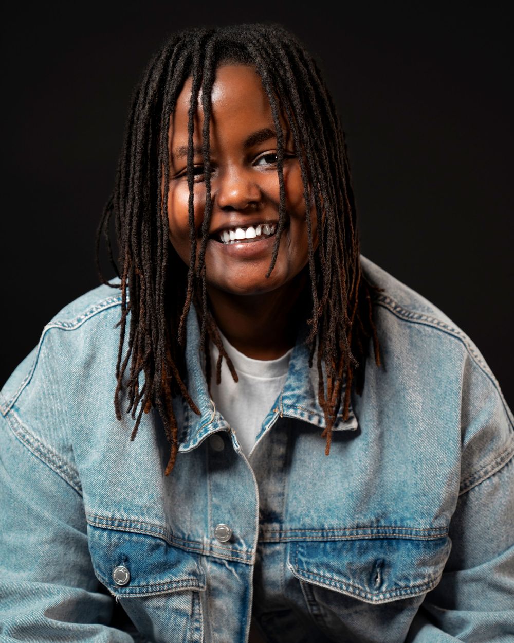 Young Black woman with dreadlocks smiles brightly wearing an oversized denim jacket against dark background.