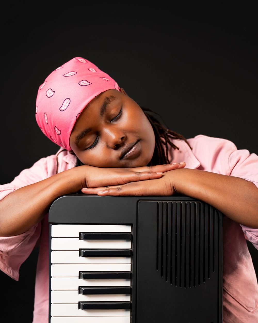 Young Black woman in pink headscarf rests peacefully on a keyboard instrument with eyes closed.