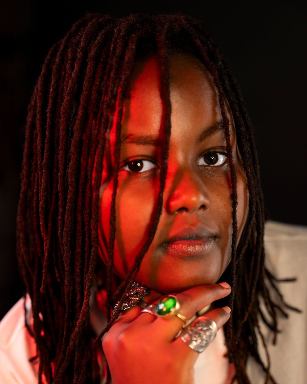 Young Black woman with red-highlighted dreadlocks poses with ringed fingers under chin in dramatic red lighting.