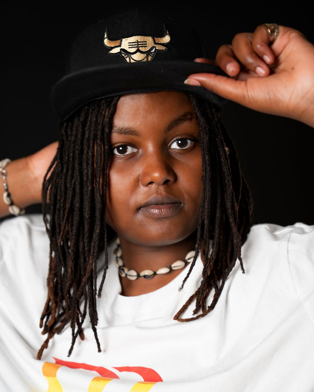 Young Black woman with dreadlocks wearing Chicago Bulls snapback hat and shell necklace poses confidently.