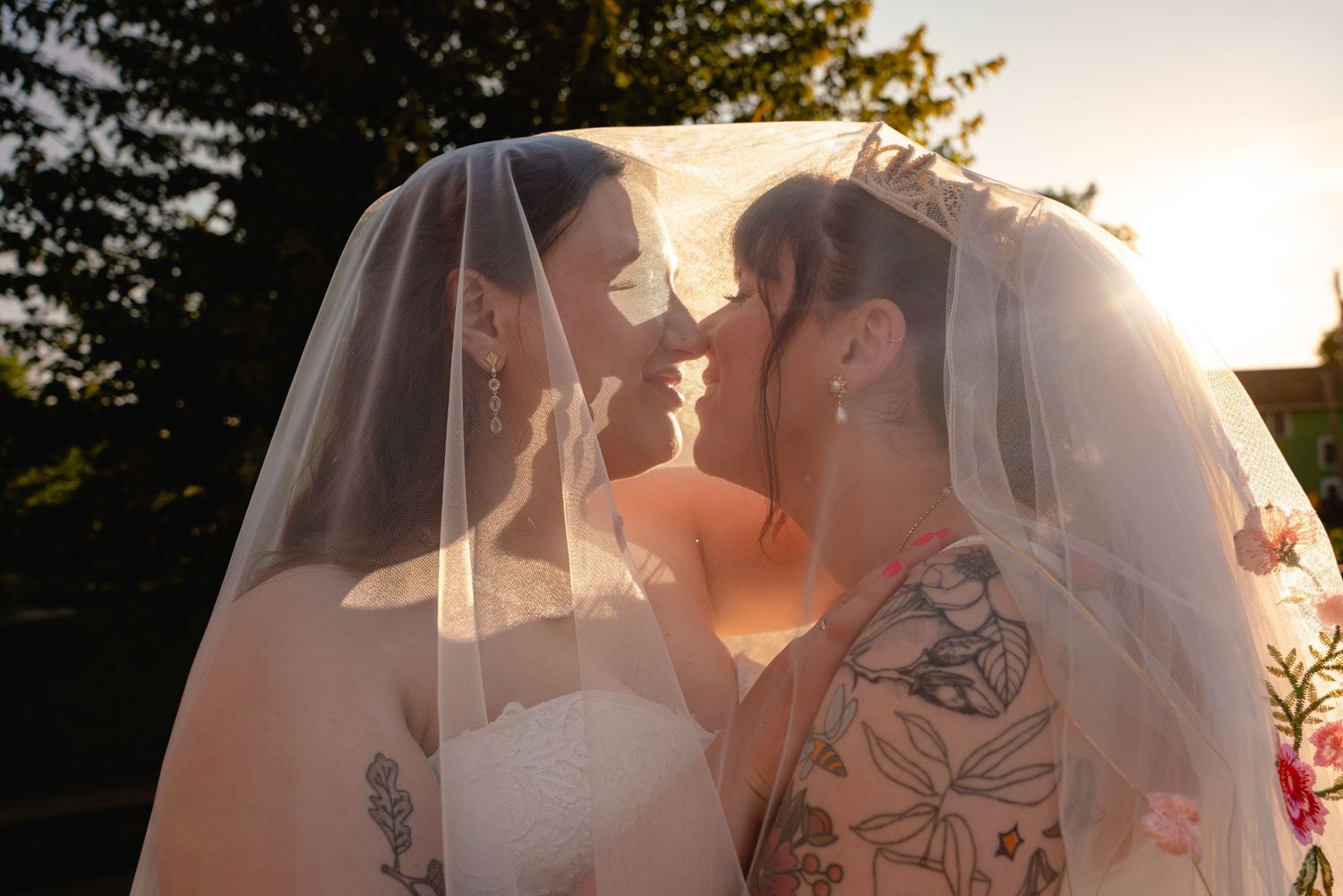 Two brides sharing a romantic moment under a veil, backlit by golden sunset light, one with floral tattoos.
