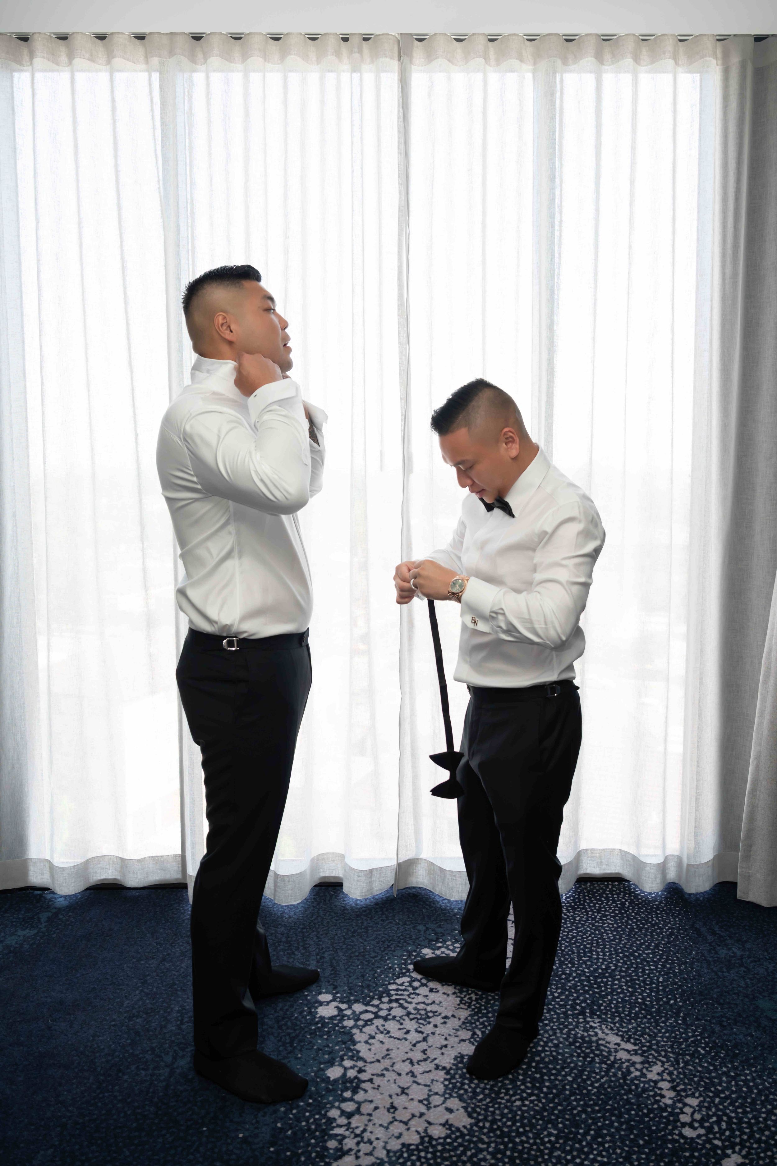Two men in white shirts and black pants getting ready, one adjusting his collar and the other holding a tie.