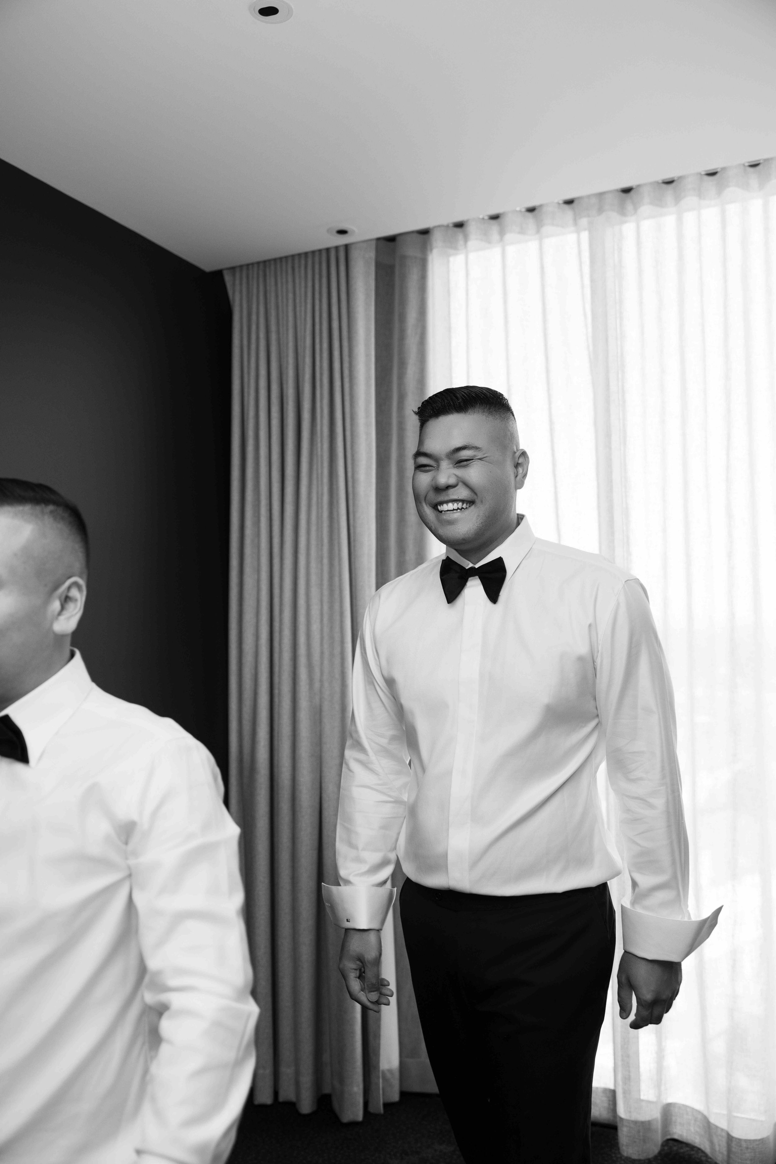 Black and white photo of smiling groom in white dress shirt and bow tie standing with groomsmen before wedding ceremony.