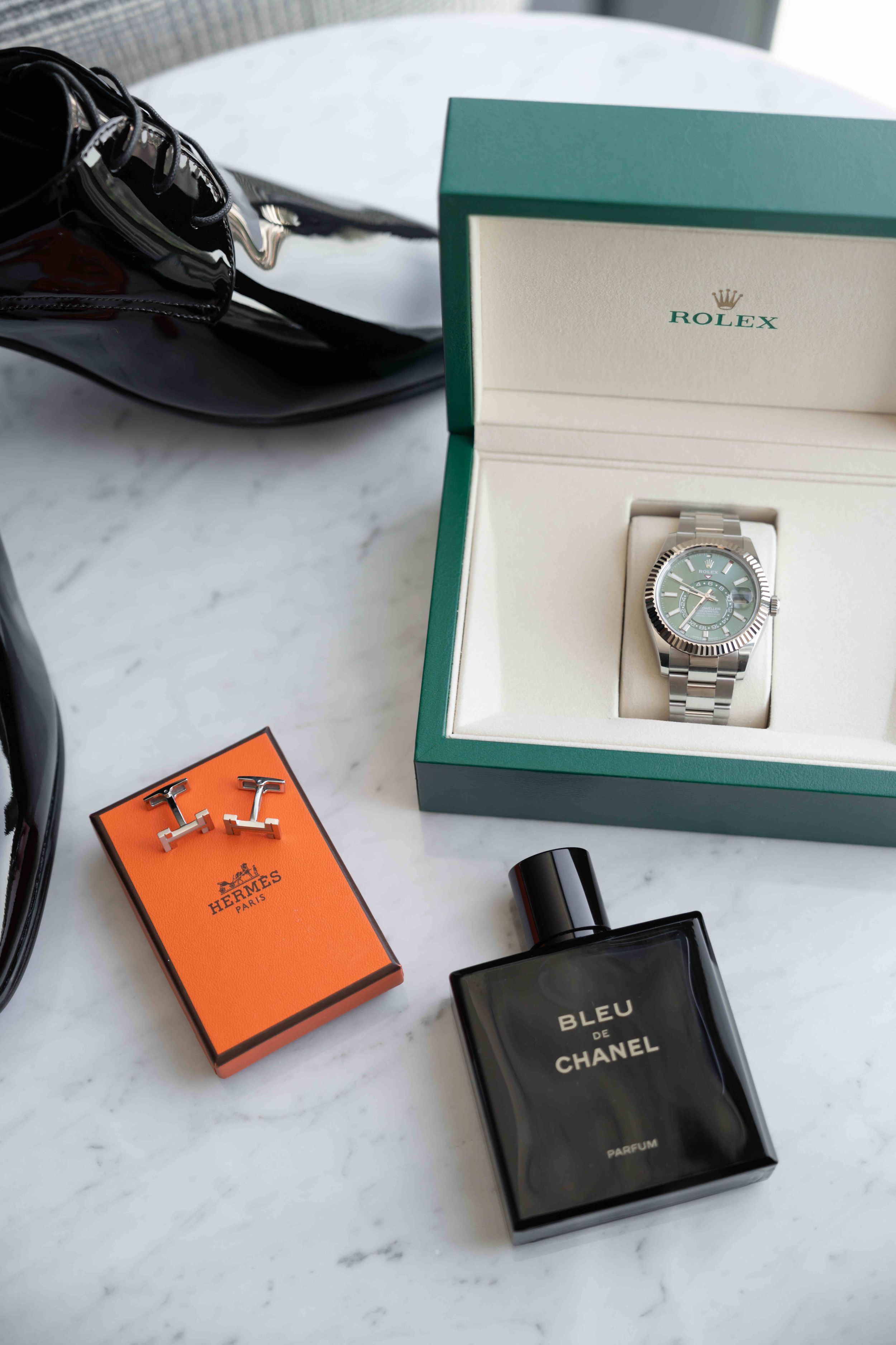 Rolex watch in green box, Hermes cufflinks, Bleu de Chanel parfum, and leather shoes on marble surface.