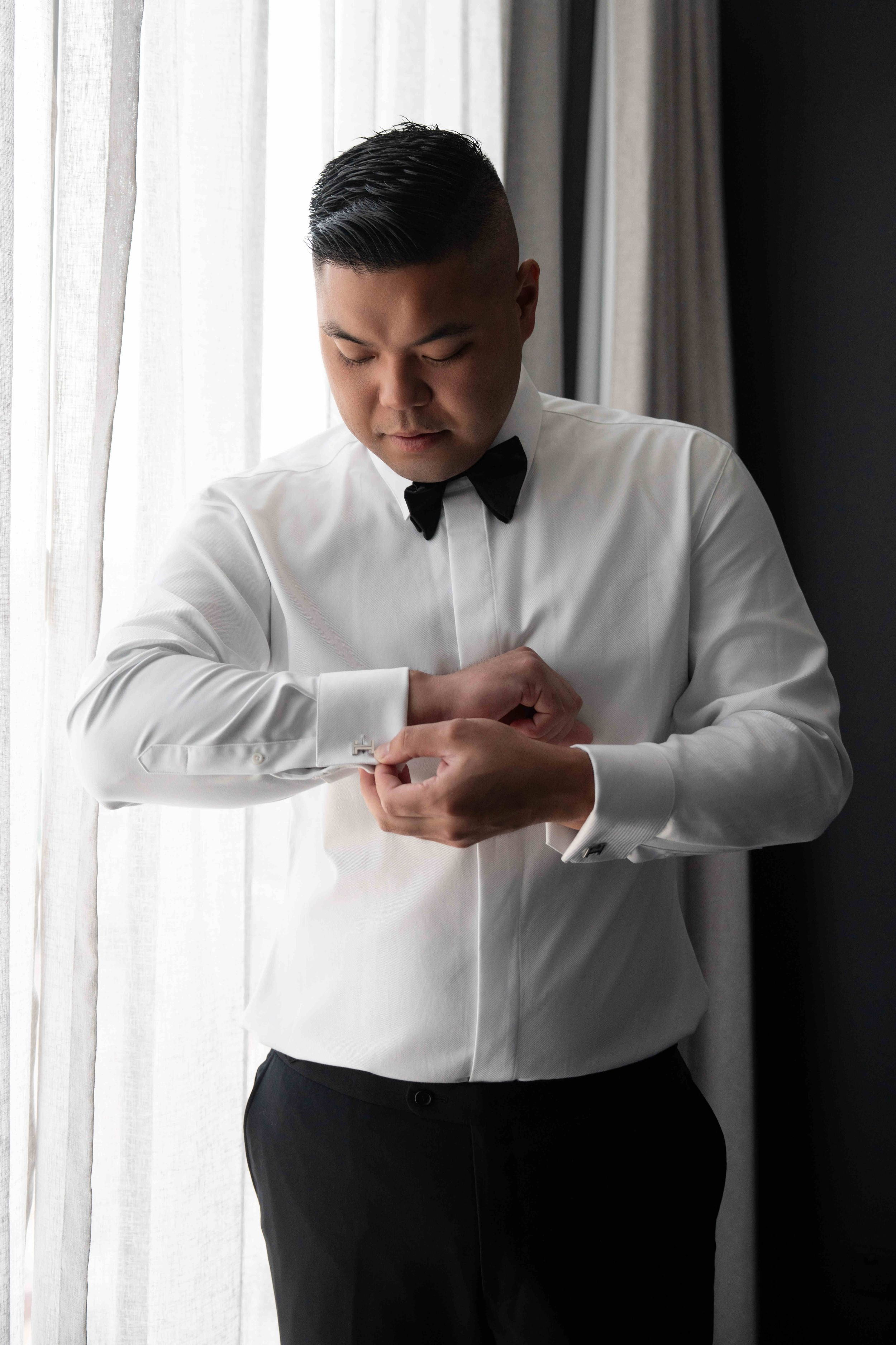 A groom in a white dress shirt and black bow tie fastening his cufflinks while standing near a window.