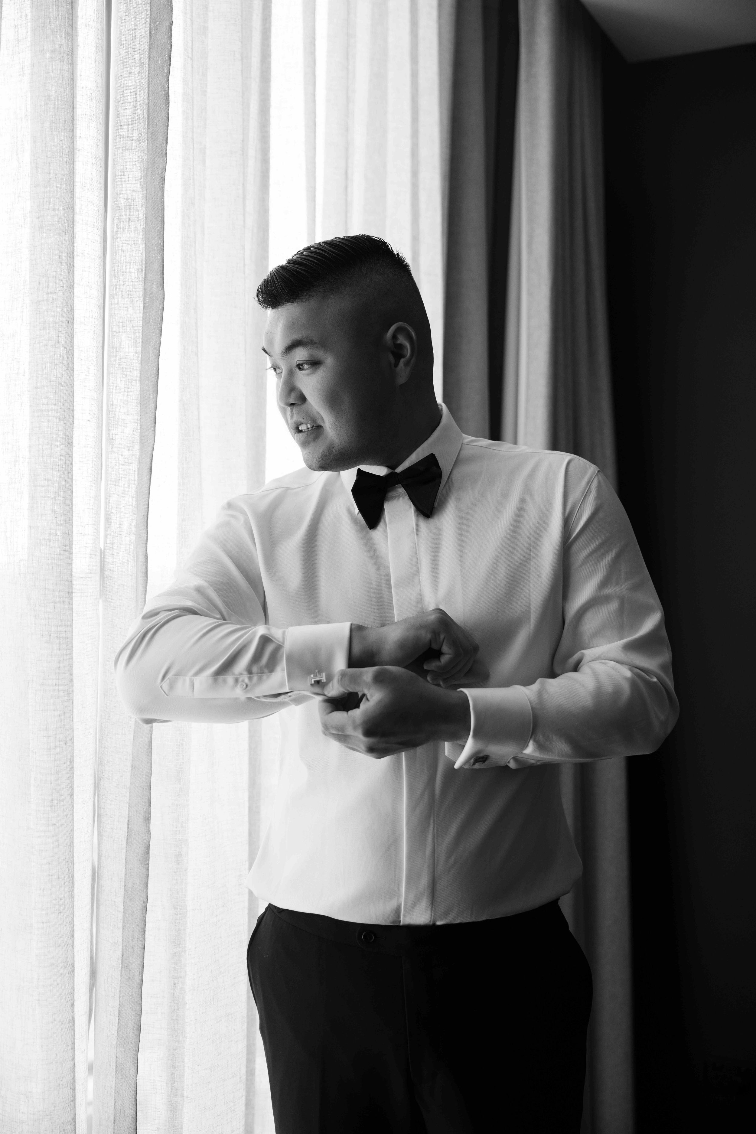 Black and white photo of a well-dressed man in a tuxedo shirt and bow tie adjusting his cufflinks by a window.