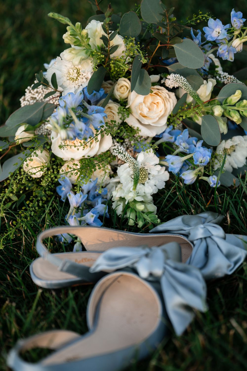 Blue and white wedding bouquet with roses and hydrangeas beside elegant light blue satin bow heels on green grass.