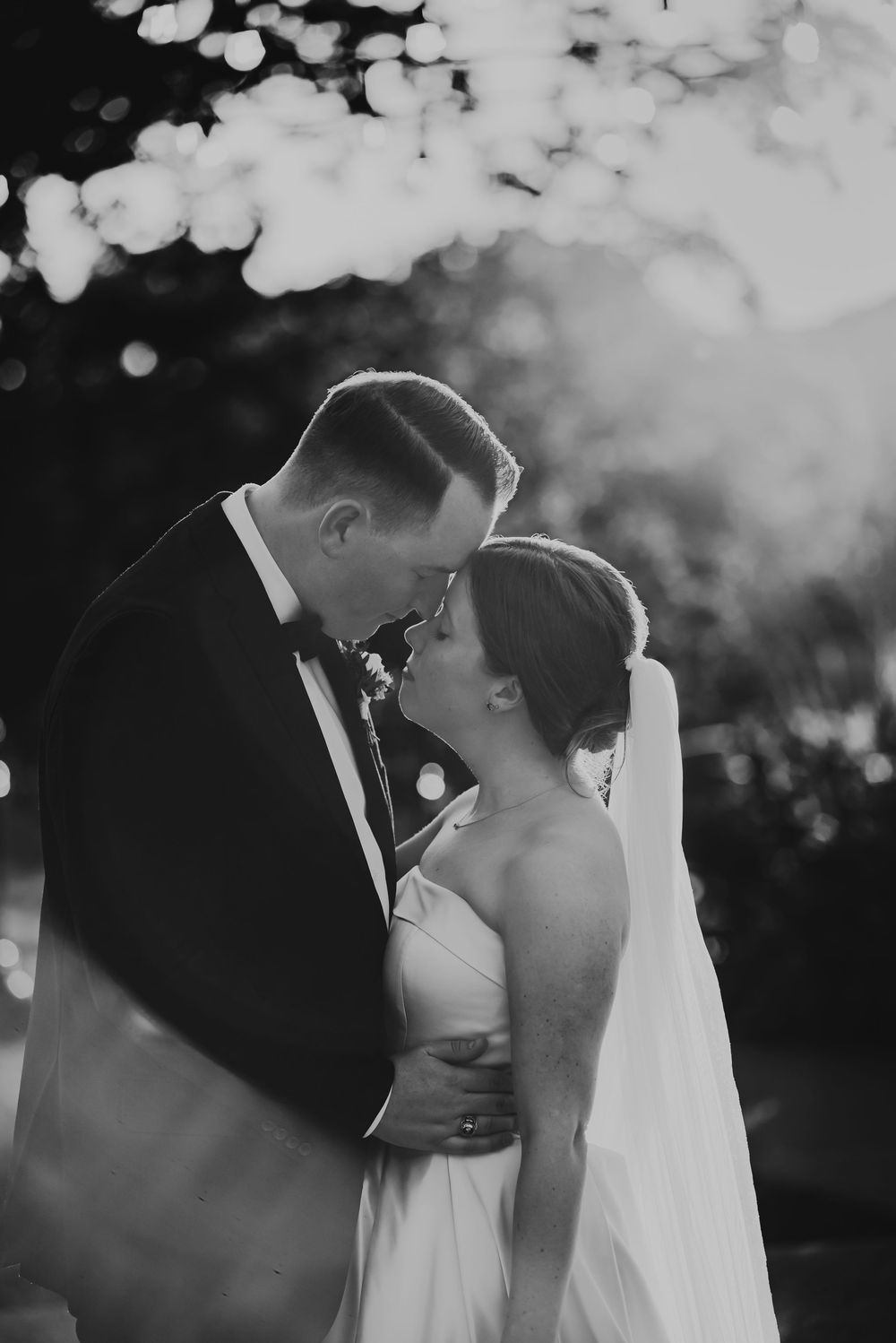 Black and white photo of a bride and groom touching foreheads, sharing an intimate moment outdoors with bokeh lights.