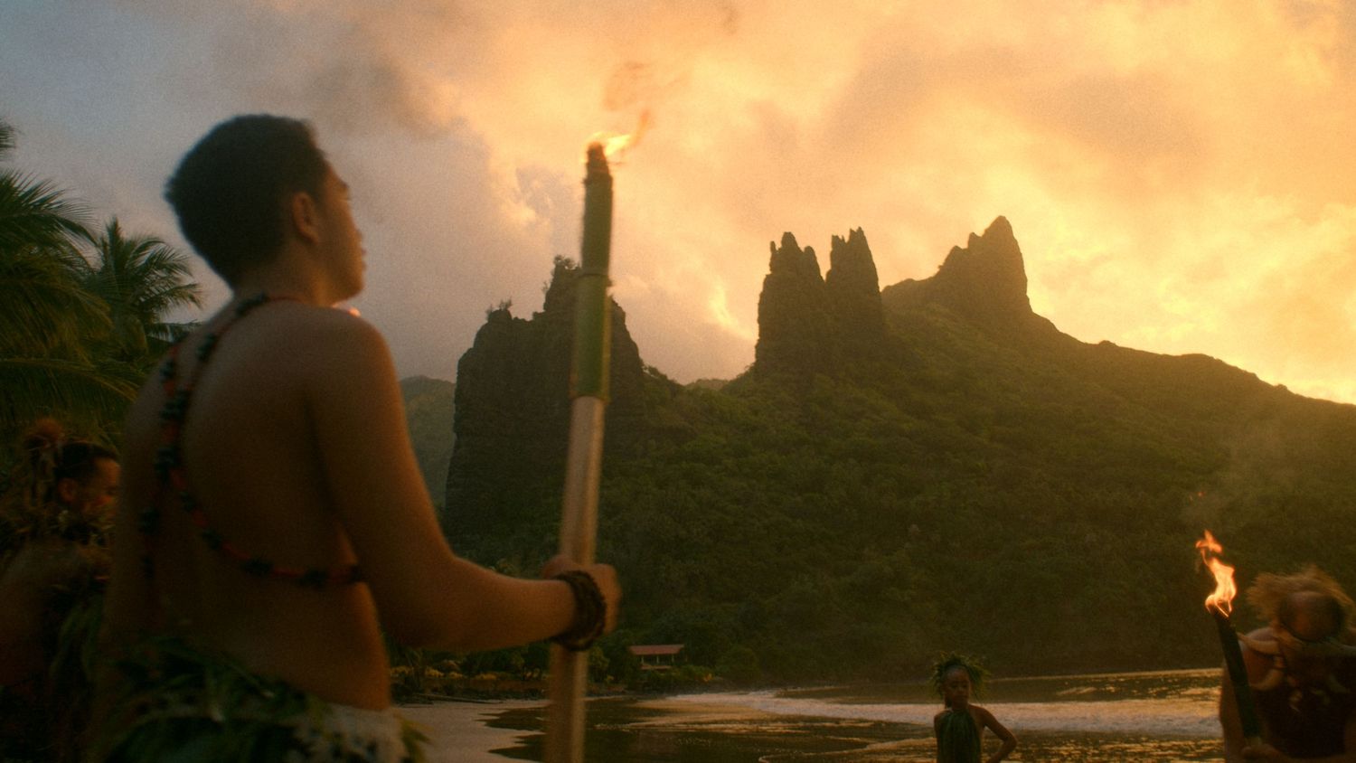 A warrior holds a spear overlooking a dramatic volcanic island landscape at sunset with torches burning nearby.