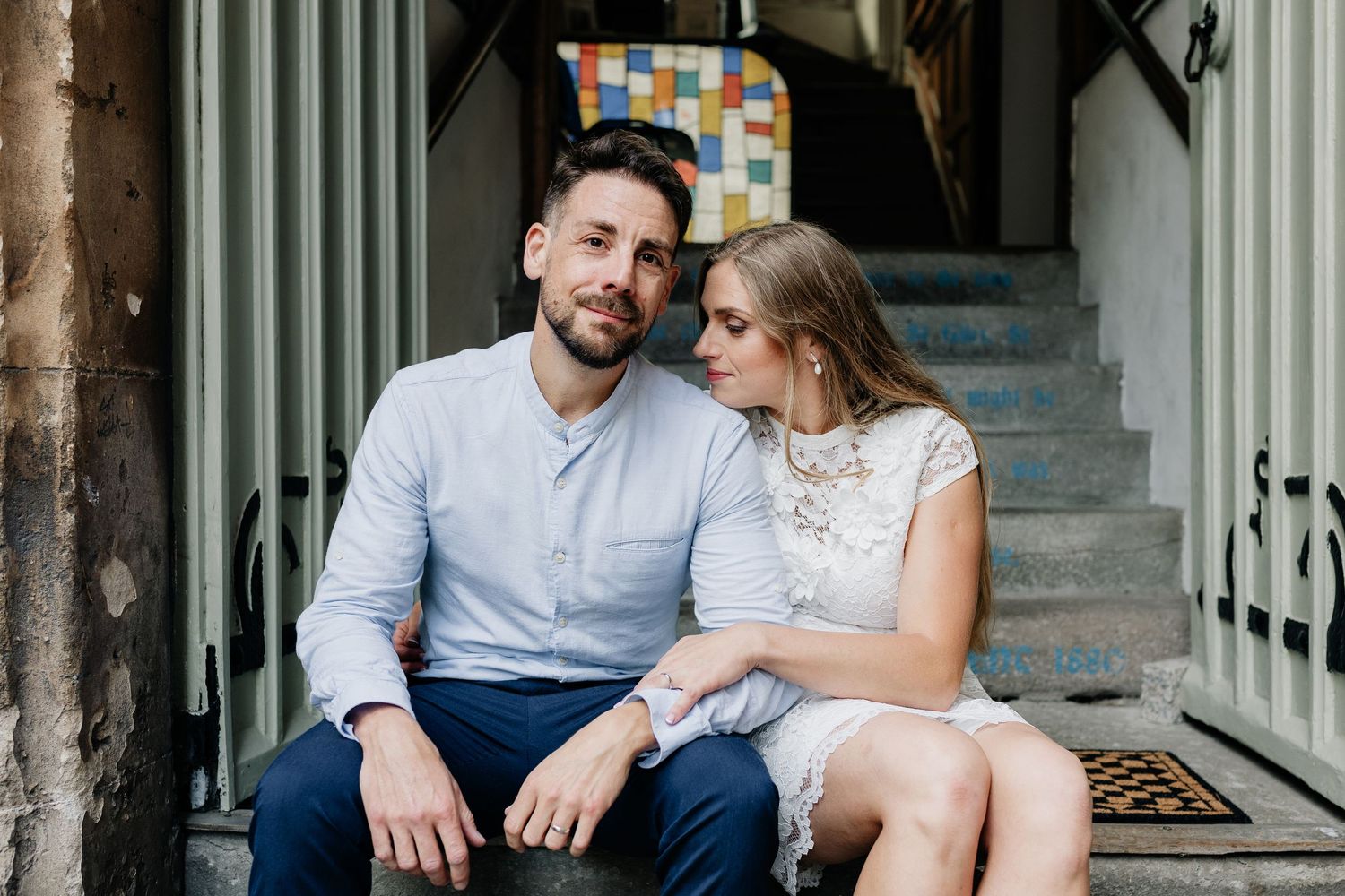 A couple sits together on stone steps, the man in a blue shirt and woman in a white lace dress, sharing a tender moment.