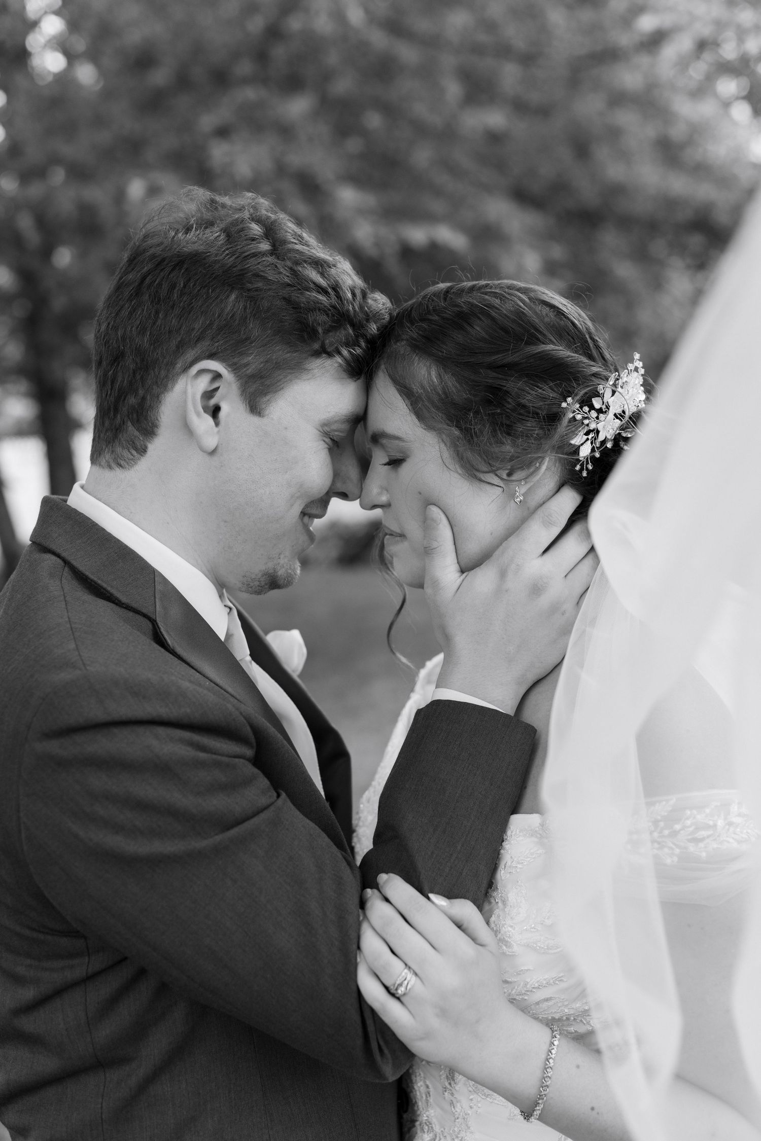 Black and white photo of a groom tenderly cupping his bride's face as they touch foreheads on their wedding day.