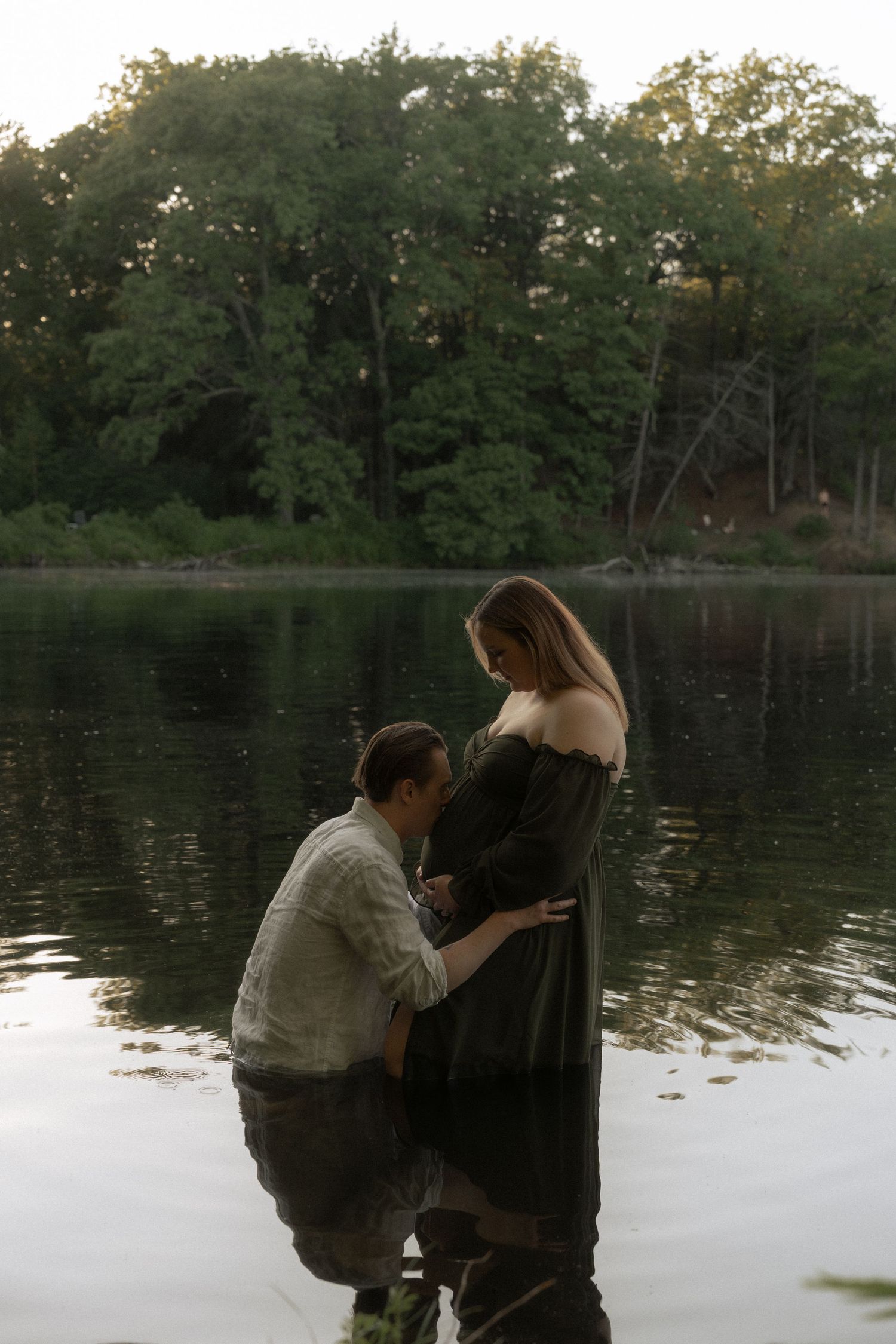 A man kneels in a lake touching a pregnant woman's belly, surrounded by lush green trees at dusk.
