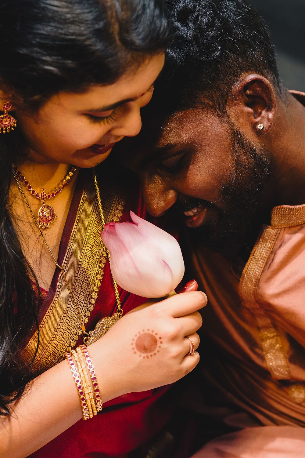 Indian couple in traditional attire sharing a tender moment with a pink lotus flower, bride wearing henna and gold jewelry.