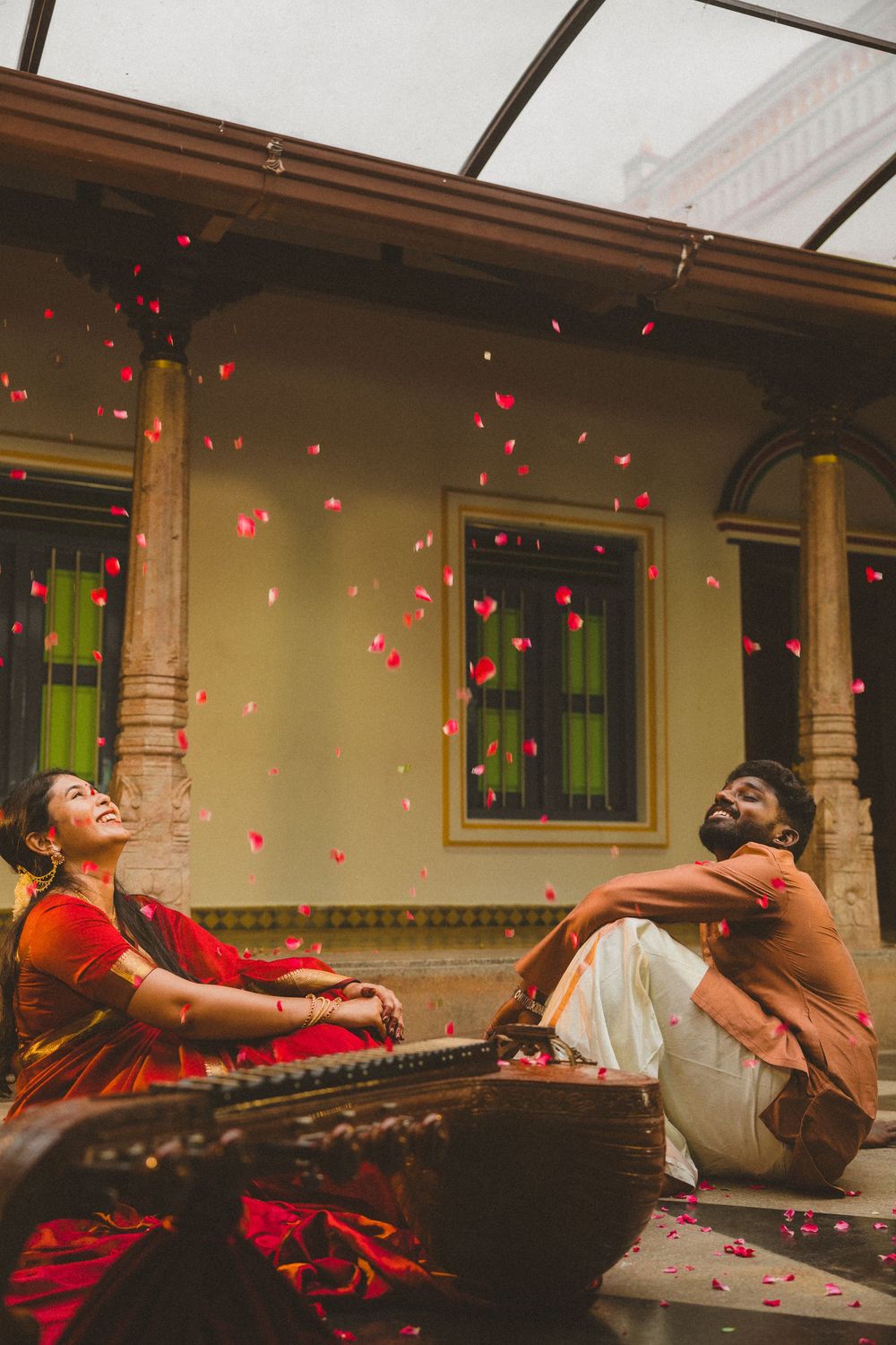 A joyful couple in traditional Indian attire laughs as red rose petals fall around them at a heritage building.