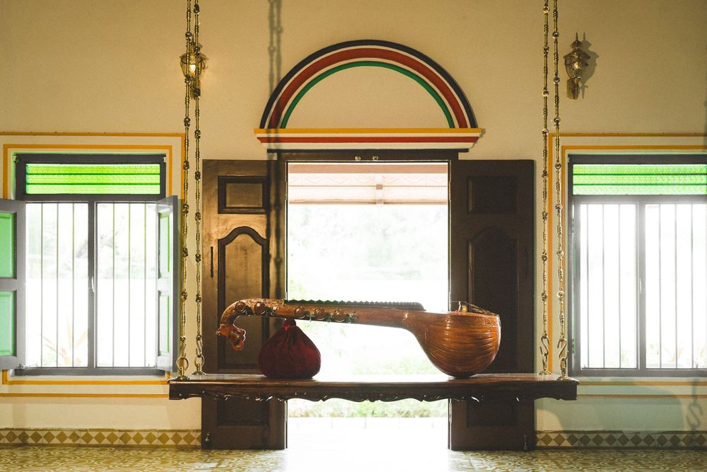 A veena instrument resting on a wooden swing inside a colorful traditional Indian room with arched decor.