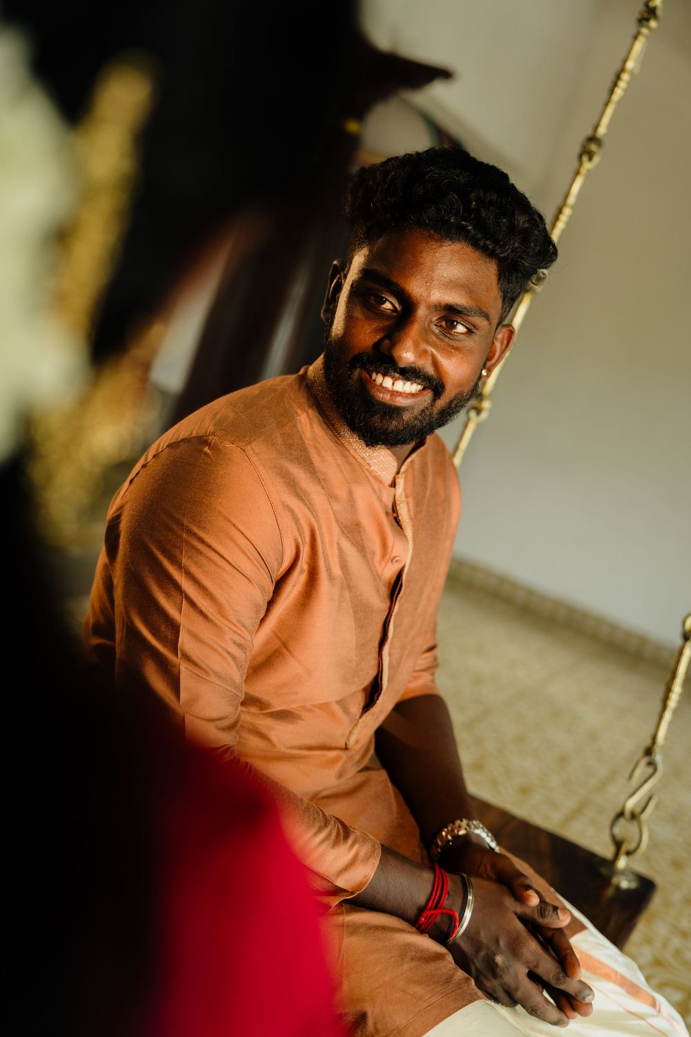 Smiling young man in orange kurta sitting relaxed indoors, wearing bracelets and looking cheerfully to the side.