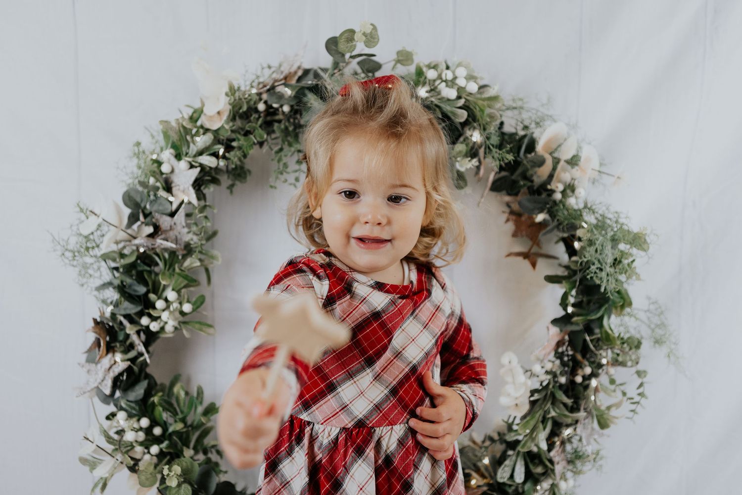 A smiling toddler girl in a red plaid dress poses inside a festive green Christmas wreath with white flowers for a Cornwall mini photo shoot