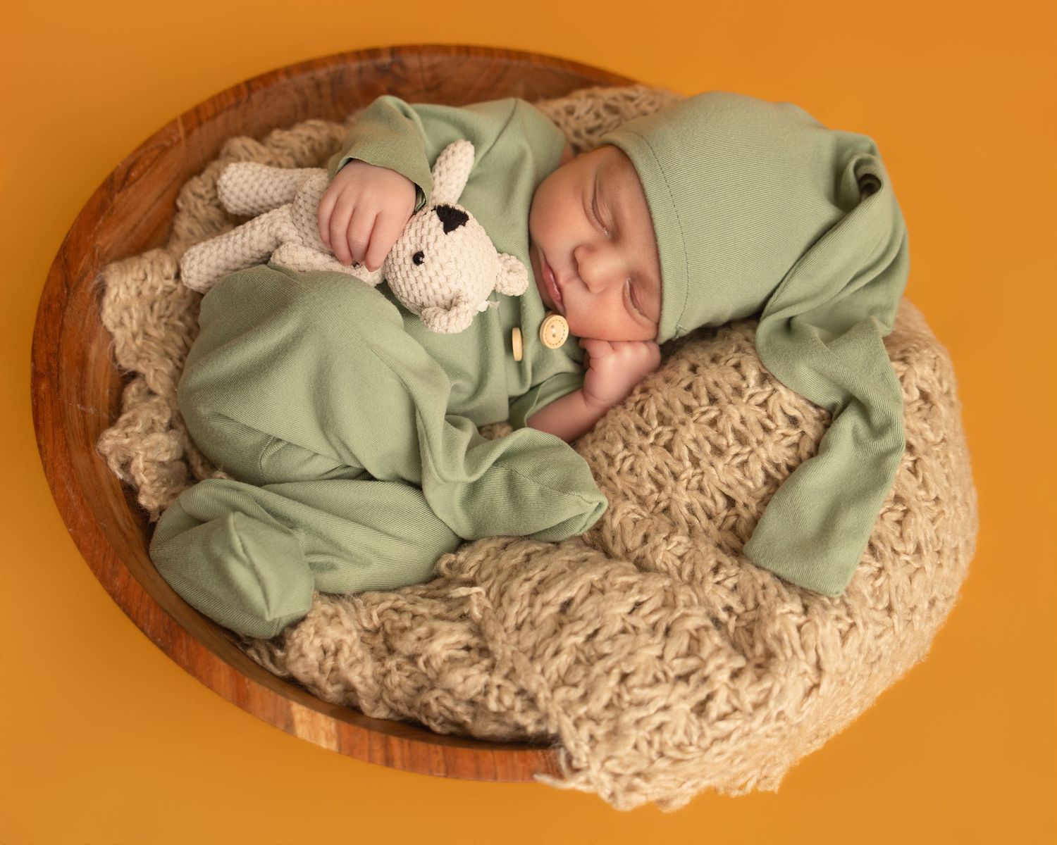 Newborn baby sleeping in wooden bowl wearing green outfit with matching hat, cuddling a knit bunny toy on beige blanket.