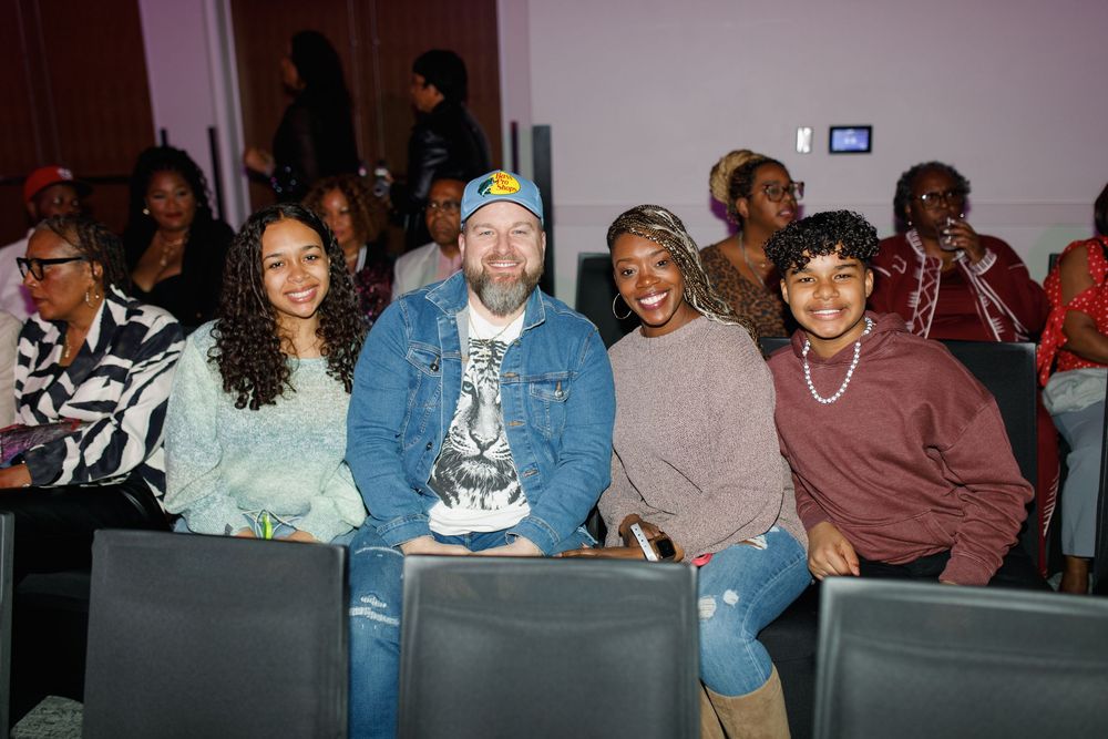 Four smiling people pose together in auditorium seats at an indoor event or show.