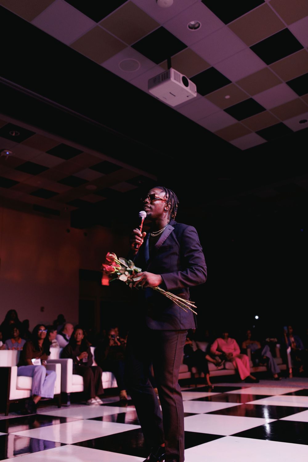 Man in suit holding flowers and microphone speaks on checkered dance floor at elegant event.