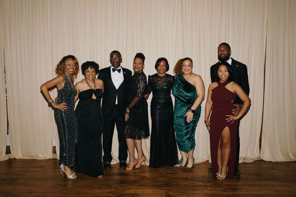 Group of eight elegantly dressed adults posing together at a formal event in front of a white curtain backdrop.