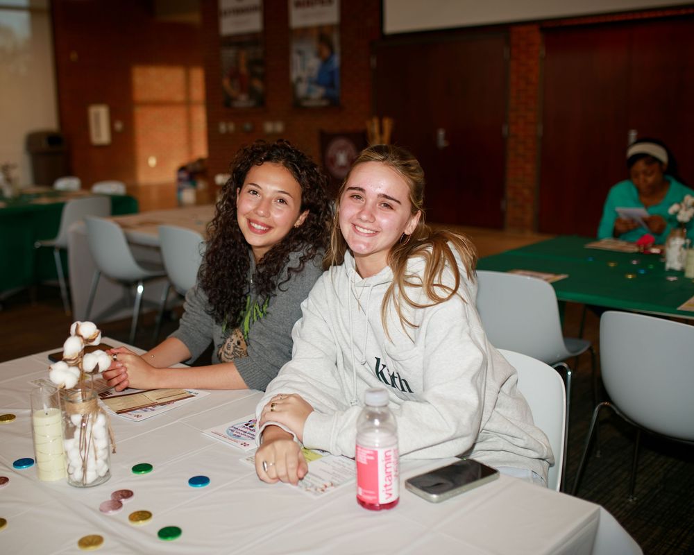 Two smiling teenage girls sitting together at a decorated table during a school event or activity.