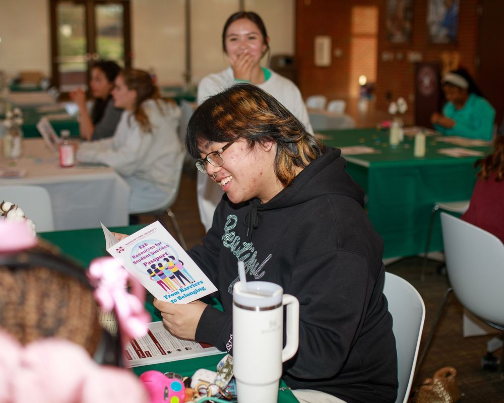 Woman smiling while reading a colorful booklet at a community event with green-themed tables in background.