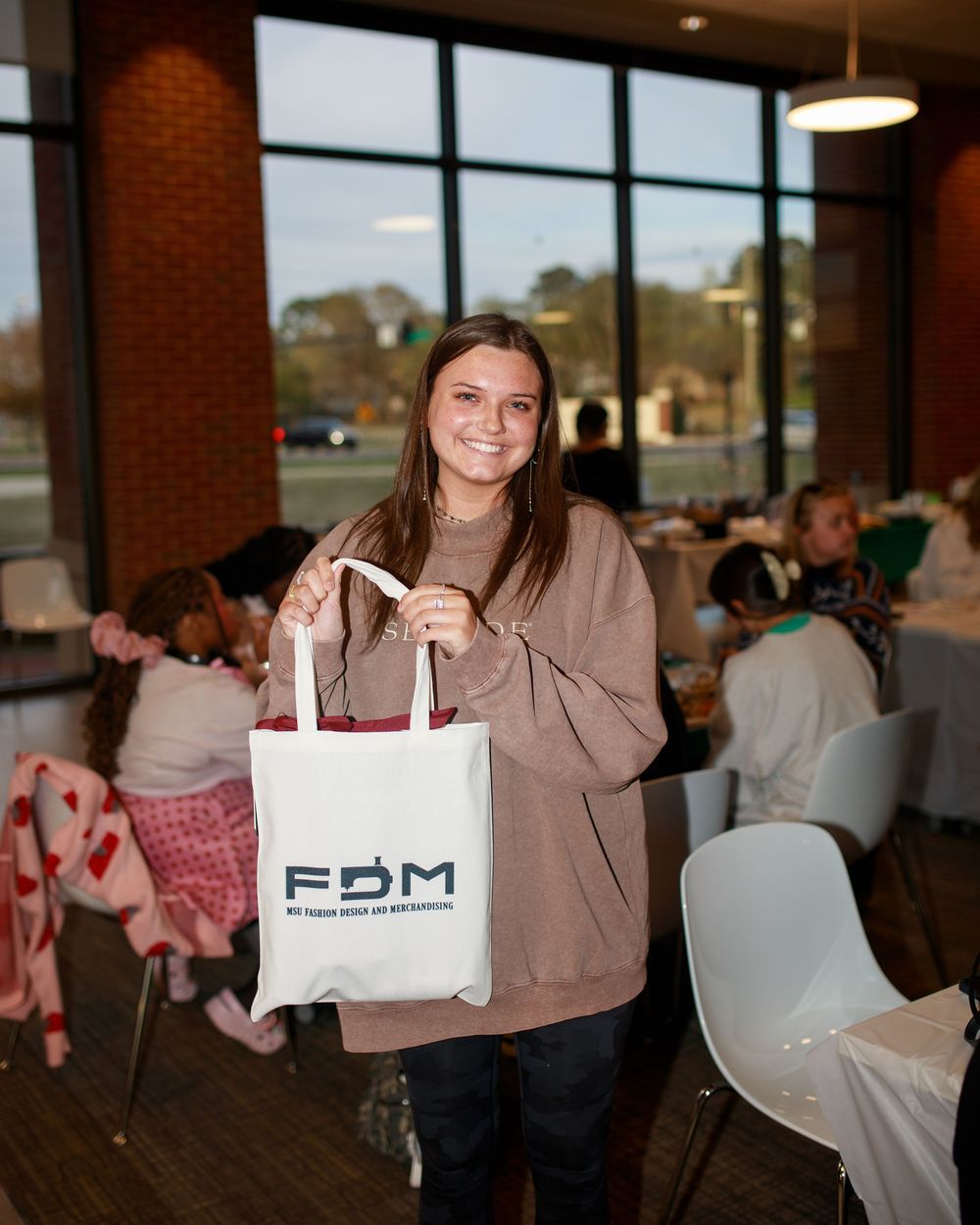 A smiling young woman holds a white FDM tote bag in a casual restaurant setting.