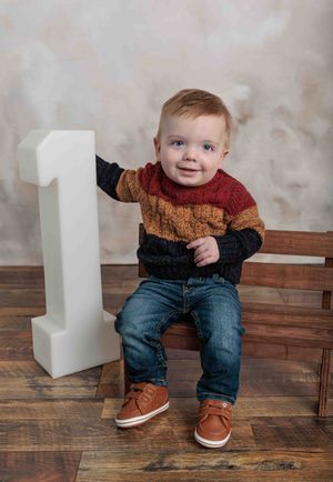 Smiling one-year-old boy in colorful striped sweater and jeans sitting beside large number one prop.