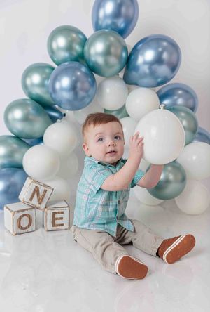 Baby boy sitting with blue and white balloons and wooden name blocks spelling NOE at first birthday party.