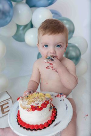 Blue-eyed baby boy eating first birthday cake at cake smash session with blue and white balloon backdrop.