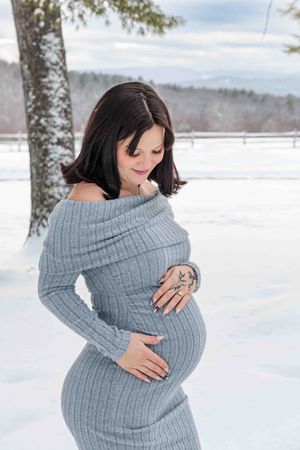 Pregnant woman in gray off-shoulder dress cradling baby bump in a snowy winter landscape.