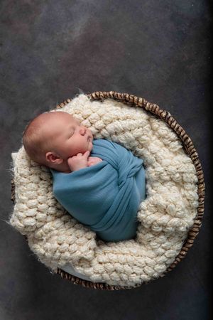 Newborn baby swaddled in blue wrap sleeping in a woven basket with cream knit blanket.