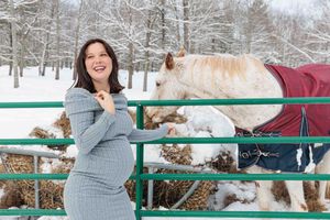 Smiling pregnant woman in grey dress posing outdoors in winter snow beside a horse wearing a red blanket.