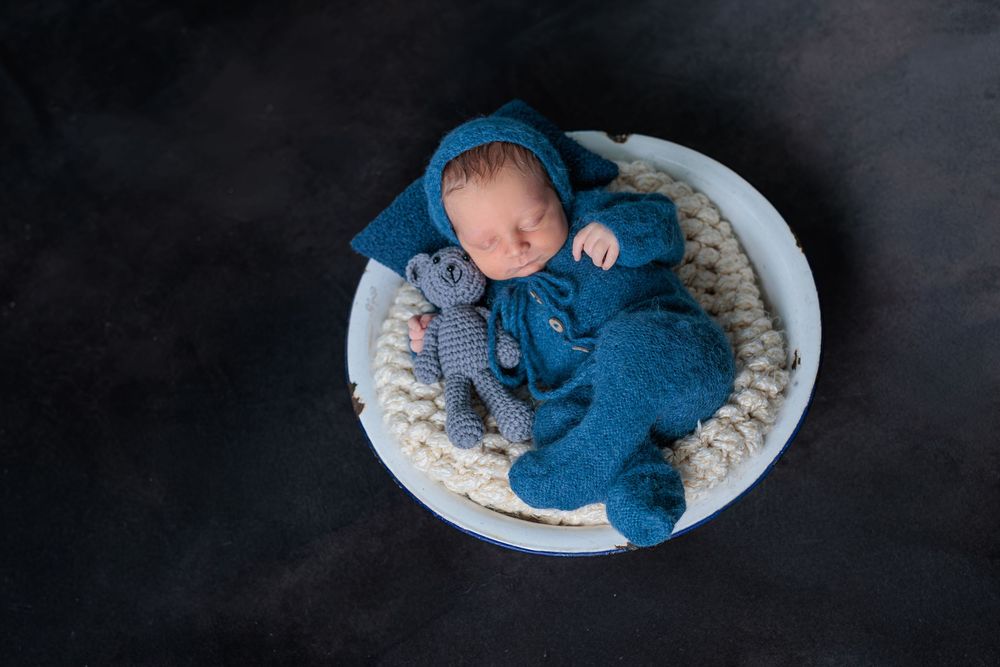 Sleeping newborn in blue knit outfit with matching hat and stuffed toy in a white bowl.