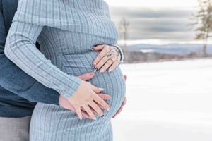 Pregnant woman in grey dress holding baby bump with partner embracing her outdoors in snowy winter landscape.