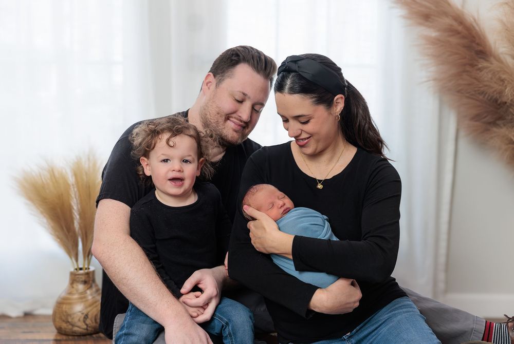 Happy family of four in black outfits posing with newborn baby in bright studio.