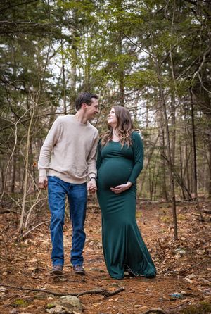 Couple holding hands on a forest path, smiling at each other during a maternity photoshoot.