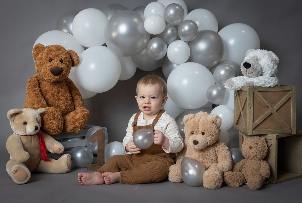 Adorable baby surrounded by teddy bears and silver balloons on gray background.