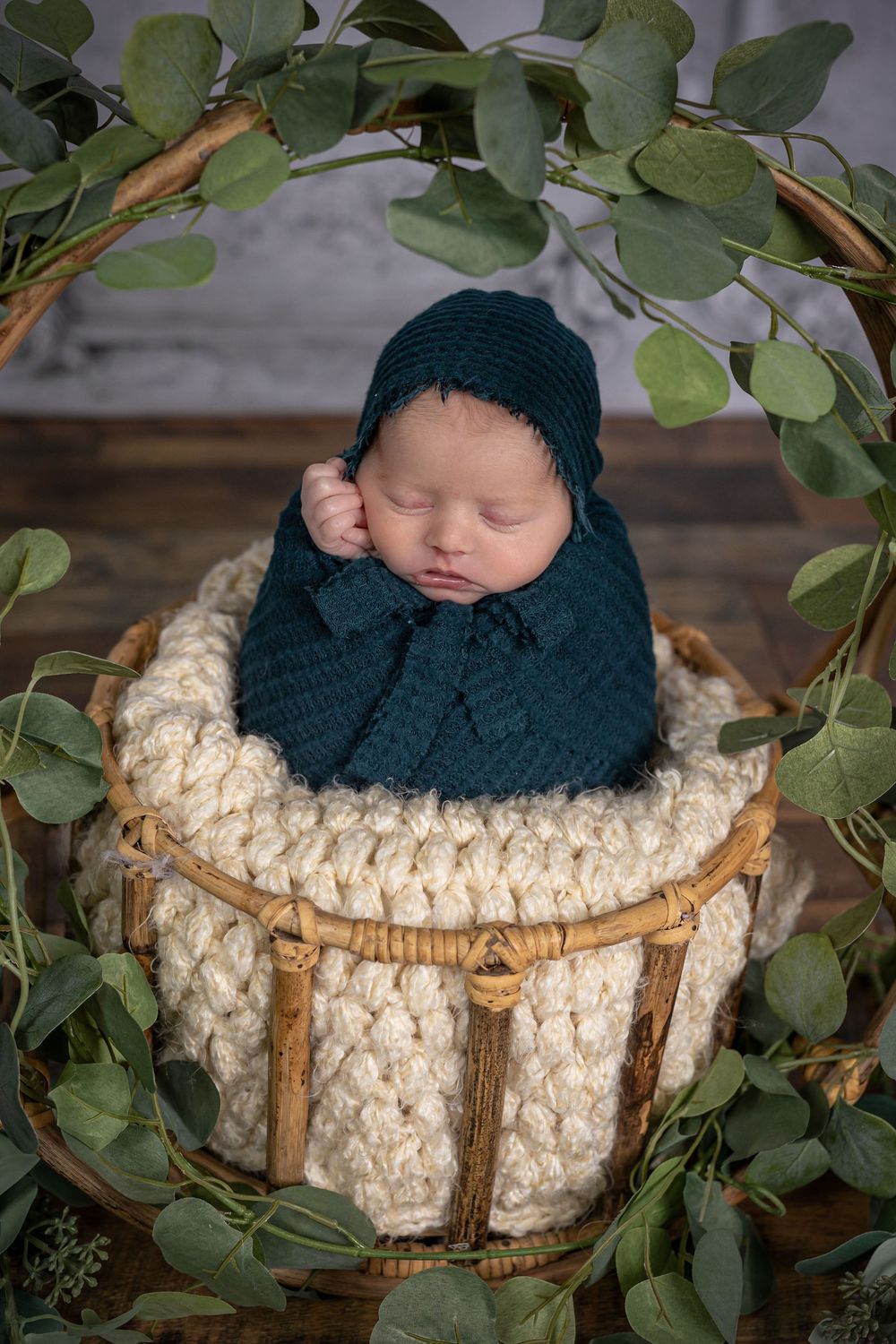 Sleeping newborn wrapped in teal knit blanket and bonnet in a wicker basket with greenery.