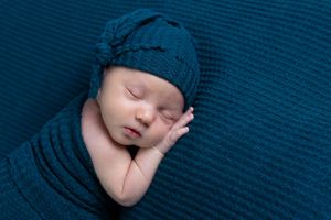 Newborn baby swaddled in teal blue wrap and matching knit hat sleeping peacefully.