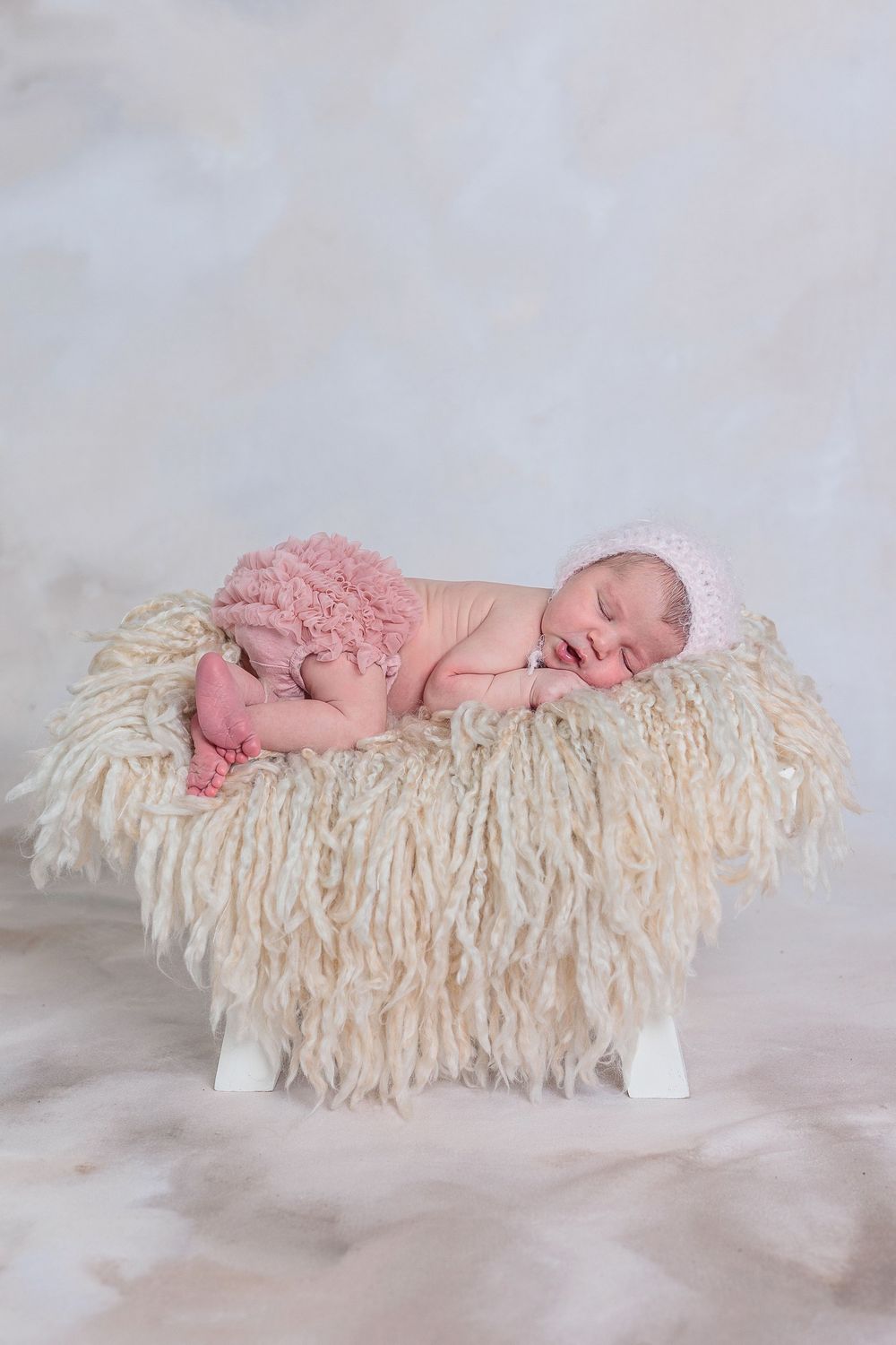 Sleeping newborn baby girl in pink bonnet resting on fluffy cream textured surface.
