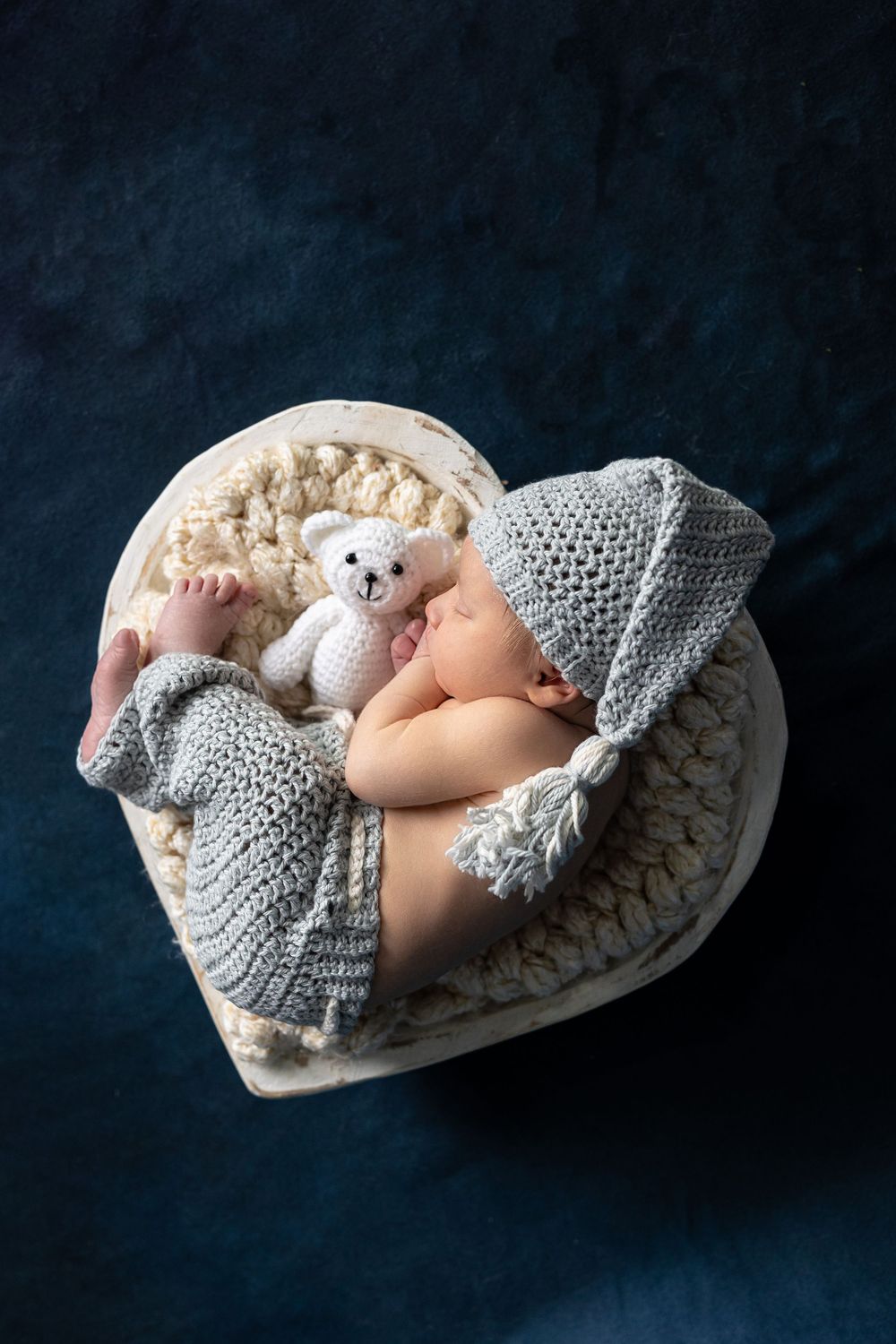 Sleeping newborn baby in grey knit hat holding white teddy bear in heart-shaped basket on dark background.