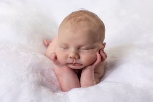 Peaceful sleeping newborn baby resting chin on hands on a soft white fluffy blanket.