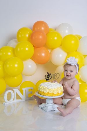 Happy baby girl in white bow headband sitting beside yellow birthday cake at ONE cake smash session.