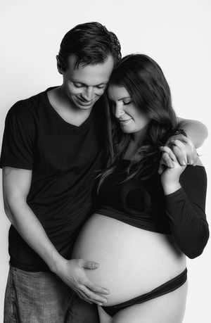 Black and white maternity photo of smiling couple in black outfits tenderly cradling pregnant belly.