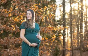 Pregnant woman in green dress cradling baby bump among autumn foliage in a sunlit forest.