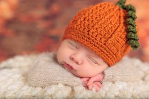 Sleeping newborn wearing an orange crocheted pumpkin hat on a soft cream blanket.