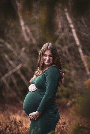 Smiling pregnant woman in green dress cradling baby bump outdoors in autumn forest setting.