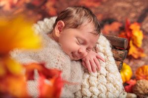 Smiling sleeping newborn wrapped in cream knit blanket surrounded by autumn leaves decor.